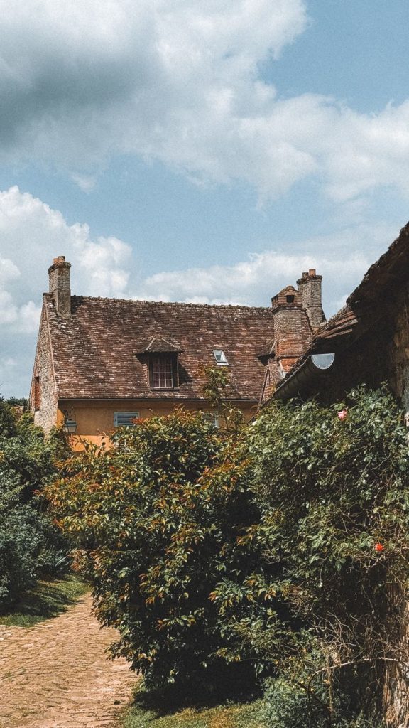 A quaint rustic cottage surrounded by lush greenery under a bright sky, capturing a tranquil village setting.